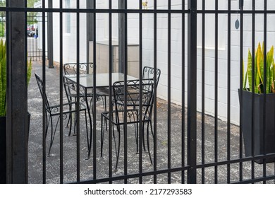 Multiple Empty Black Metal Tables And Chairs On A Cafe Sidewalk Patio Of A Restaurant. The Exterior Brick Wall And Background Are White And There's A Black Fence And Gate With Potted Flowers In Front.