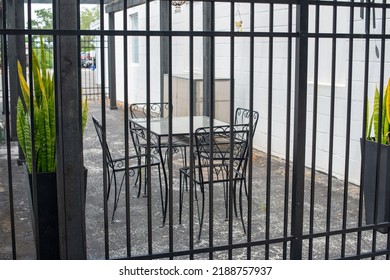 Multiple Empty Black Metal Tables And Chairs On A Cafe Sidewalk Patio Of A Restaurant. The Exterior Brick Wall And Background Are White And There's A Black Fence And Gate With Potted Flowers In Front.