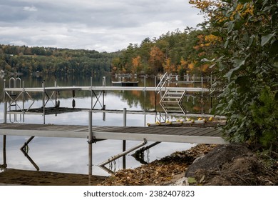 Multiple empty boat docks at the end of the summer season.  Fall foliage is in the background.  There is calm water with reflections.