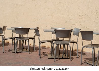 Multiple Empty Grey Metal Tables And Chairs On A Cafe Sidewalk Patio Of A Restaurant. The Exterior Brick Wall And Background Are Yellow Colored With A Pink And Gray Colored Tile On The Ground.  