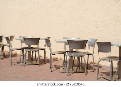 Multiple Empty Grey Metal Tables And Chairs On A Cafe Sidewalk Patio Of A Restaurant. The Exterior Brick Wall And Background Are Yellow Colored With A Pink And Gray Colored Tile On The Ground.  