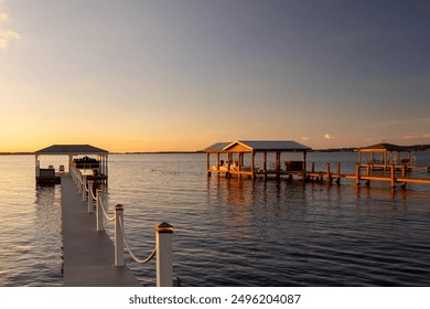 Multiple floating docks with custom wooden shelters, awnings and lifts for protecting recreational boats. The lake's water is calm with a reflective golden color from the glow of the setting sun.  - Powered by Shutterstock