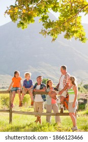 Multiple Generations Of A Family With Backpacks Resting By The Fence Of A Field At The Countryside.