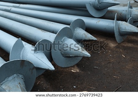Multiple gray painted helical piers lying on the ground at a construction site. Long steel screw piles with pointed tips and blades prepared for building foundation installation.