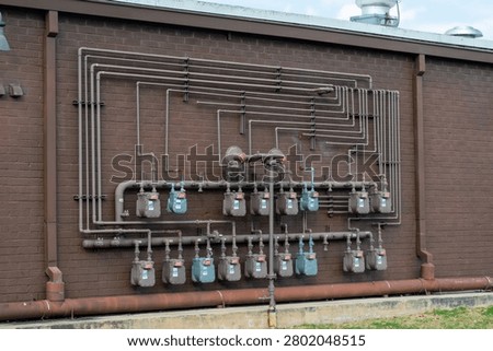 Multiple grey gas meters and interconnected utility pipes distributing natural gas for an urban building against a textured brown brick wall exterior, representing essential infrastructure and energy supply.