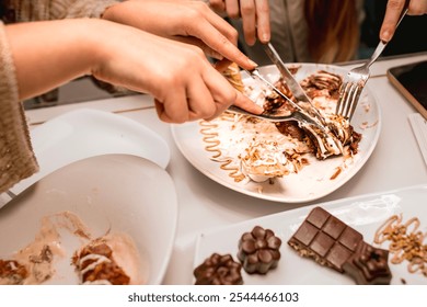 Multiple hands eagerly sharing a gourmet pancake dessert topped with white chocolate and pistachios, using forks and knives on a partially eaten plate. Chocolate treats accompany the scene - Powered by Shutterstock