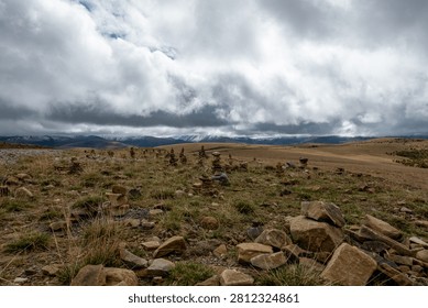 Multiple hand-stacked stone cairns dot a vast golden-brown highland steppe plateau in the Altai Republic of southern Siberia, Russia, with snow-dusted mountain ridges stretching across the horizon. Heavy dramatic storm clouds dominate the sky, casting moody diffused light across the open grassland and lending the scene a raw, atmospheric quality characteristic of this remote region. Available for editorial and commercial licensing.