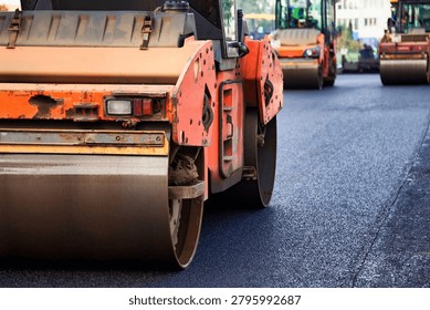 Multiple industrial road rollers compact fresh asphalt concrete on a wide street during urban construction work.