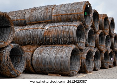 Multiple large rolls of rust-colored steel wire mesh stacked outdoors against a cloudy sky backdrop, showcasing industrial strength and texture.