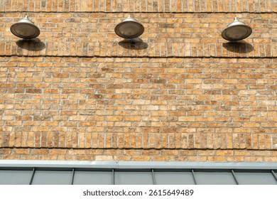 Multiple light grey colored barn lights, in a row, on the outside of an old yellowish beige brick building. The three light fixtures have silver shades with flood lights directed downwards but off. 