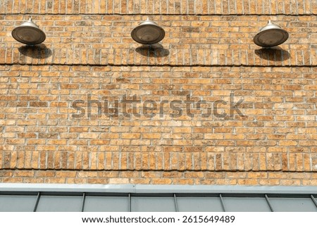 Multiple light grey colored barn lights, in a row, on the outside of an old yellowish beige brick building. The three light fixtures have silver shades with flood lights directed downwards but off. 