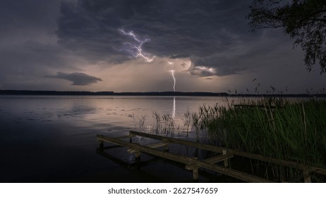 Multiple lightning bolts strike over a calm lake during a thunderstorm in Masuria, Poland. Old wooden pier in the foreground.