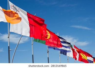 Multiple national flags flutter in the wind under a bright blue sky, representing different countries with unique colors and symbols