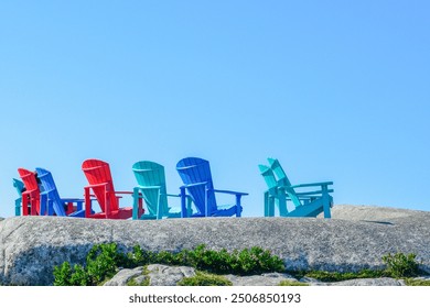 Multiple outdoor blue, red, and teal green polywood recycled plastic Adirondack chairs on a rocky cliff in a row overlooking the ocean. The sky is bright blue and the ground has patches of grass.  - Powered by Shutterstock