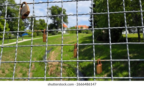 Multiple padlocks hang on grid-pattern fence, symbolizing love and commitment, with walking paths, trees, and lawns visible through the wire in vibrant urban park