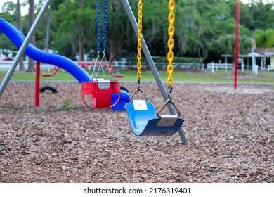 Multiple Plastic And Rubber Swings Hanging From Chains In A Children's Park. There's A Blue Plastic Slide In The Background With Large Lush Trees And Benches. The Ground Is Covered In Tree Mulch. 