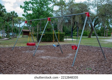 Multiple Plastic And Rubber Swings Hanging From Chains In A Children's Park. There's A Blue Plastic Slide In The Background With Large Lush Trees And Benches. The Ground Is Covered In Tree Mulch. 