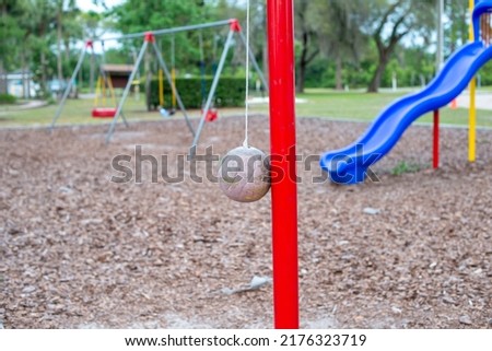 Multiple plastic and rubber swings hanging from chains in a children's park. There's a blue plastic slide in the background with large lush trees and benches. The ground is covered in tree mulch. 
