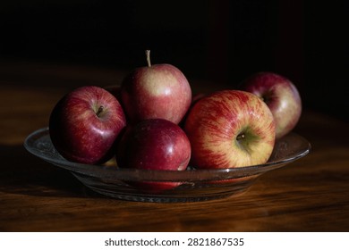 Multiple red, crispy apples on a clear glass plate in the center of a wooden table. The small crab apples are of various sizes and bright red and yellow, with stems attached. The wooden table has a dark woodgrain. 