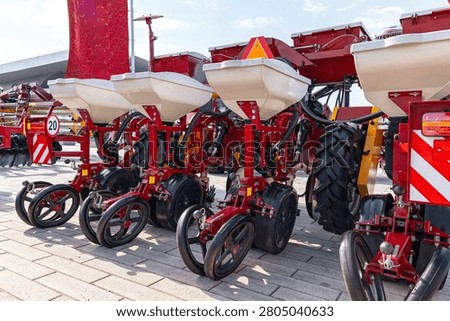 Multiple red tractor-mounted seed planters lined up outdoors on a paved surface. Visible are white seed hoppers, metal frames, black rubber wheels and coulters
