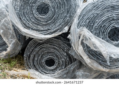 Multiple rolls of galvanized wire mesh fencing, partially covered in plastic, stacked outdoors, illustrating a durable and versatile material for construction, agriculture, or security