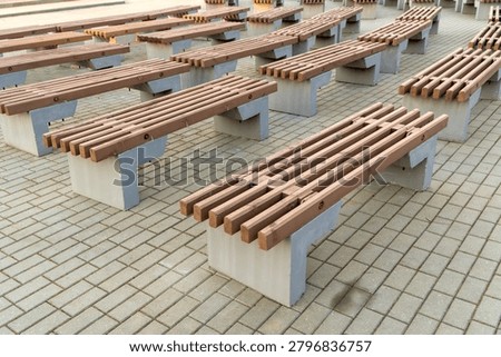 Multiple rows of empty modern wooden benches with concrete bases are arranged on a stone-paved public square or outdoor theater area.