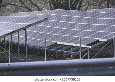 Multiple rows of solar panels are installed behind a metal mesh fence. The panels align neatly, facing the same way, while trees form a natural backdrop beyond the fenced energy installation.