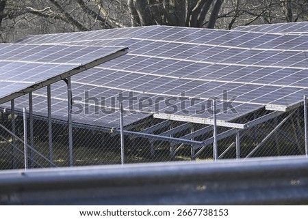 Multiple rows of solar panels are installed behind a metal mesh fence. The panels align neatly, facing the same way, while trees form a natural backdrop beyond the fenced energy installation.