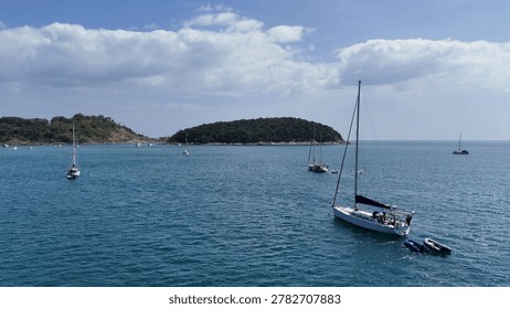 Multiple sailboats float on calm blue water near lush tropical islands under cloudy skies.