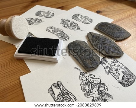 A multiple sheets of paper with black and white prints on them, beneath the hand-carved stamps used for the printing, an inkpad and a barren with felt surface.