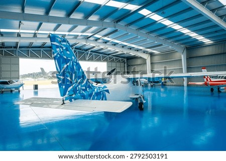 Multiple small aircraft parked inside a spacious airplane hangar featuring a striking blue epoxy floor and a geometric patterned tailfin, displaying readiness for flight and maintenance