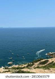 Multiple small boats navigate calm ocean waters near rocky shoreline under clear blue sky, viewed from elevated cliffside vantage point.