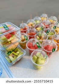 Multiple small clear plastic cups and containers filled with precut watermelon, cantaloupe, and pineapple chunks. Ready to eat fruit snack portions.