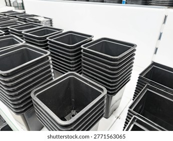 Multiple stacks of empty black square plastic plant pots arranged on white shelves in a greenhouse or nursery setting. Pots designed for seedlings, gardening, or small plants, made from durable plasti