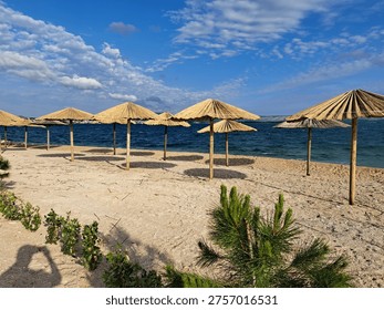 Multiple straw umbrellas lined up on a sandy beach next to the blue sea under a bright sky.
