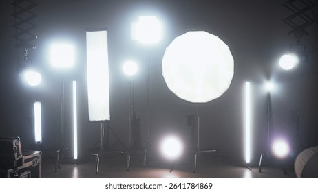 Multiple studio lights, softboxes, and LED panels illuminating a film set. Equipment includes vertical tubes, octaboxes, and overhead lighting mounts. Backstage preparation for studio professional