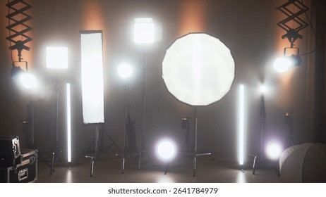 Multiple studio lights, softboxes, and LED panels illuminating a film set. Equipment includes vertical tubes, octaboxes, and overhead lighting mounts. Backstage preparation for studio professional