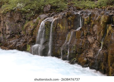 multiple thin waterfalls falling over a bare red rock into a rapid flowing mountain stream