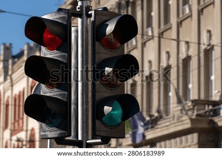 Multiple traffic lights at a city intersection with red and green signals, street infrastructure, sunlight, and blurred urban buildings in the background.