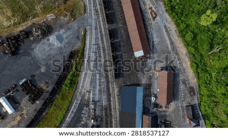 Multiple train tracks running through an industrial area. Several buildings are present alongside the tracks. The area is surrounded by trees and gravel.