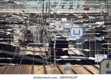 Multiple trains are on different tracks at a busy railway station. Electric cables hang above and the sun shines down on the scene. Activity is evident as trains arrive and depart
