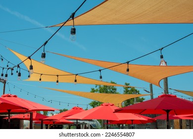 Multiple triangle shaped yellow nylon sunshades and awnings hanging over a patio deck. There are red colored canvas umbrellas hung with strings of clear patio light against a bright blue sunny sky. - Shutterstock ID 2246554807