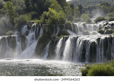 Multiple waterfalls cascade over rocky cliffs, surrounded by lush greenery and mist, creating a powerful and scenic view of nature's beauty. - Powered by Shutterstock