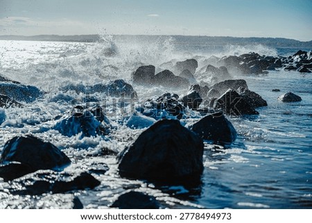 Multiple wave splashes along the rocky shoreline of New York. Wide view of sea water splashing against a line of rocks on New York coast under clear sky.