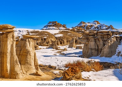 Multiple weathered hoodoo formations dot the snowy badlands landscape with distant buttes and mesas under brilliant blue sky at Bisti Badlands, New Mexico