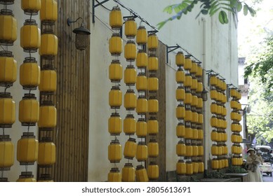 Multiple yellow lanterns hang in repeated rows along the sides of a narrow outdoor walkway. Wooden wall panels, pale building facades, greenery, and filtered daylight create a colorful decorative corridor with strong depth and rhythm.