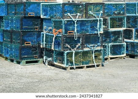 Multiple yellow lobster pots or traps stacked high along a wharf. Containers are made from yellow wire, and nylon netting, and in a square shape. There's no bait in the pots, all are empty.