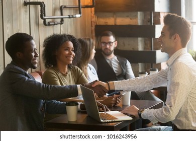 Multiracial african and caucasian men handshaking at meeting in cafe, diverse friends greeting sitting at coffeeshop table or black couple making deal shaking hands to white businessman with laptop