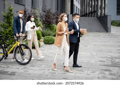 Multiracial business team in medical masks walking on city street and talking. Concept of business during Coronavirus pandemic. Asian businessman and caucasian businesswoman holding coffee and food