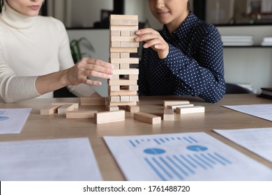 Multiracial Businesswomen Play Build Wood Block Stacking Game, Financial Stats Report On Desk Close Up. Cooperation, Company Stability, Reach Aim Effort, Business Strategic Thinking Management Concept